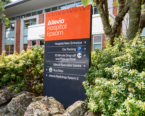 Freestanding outdoor directory sign at Allevia Hospital Epsom with arrows pointing to various hospital facilities and transport access.