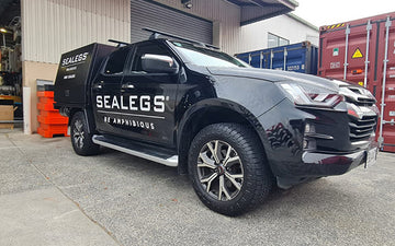 Black Sealegs utility ute featuring large white "SEALEGS®" and "BE AMPHIBIOUS" branding on the side, parked outside a workshop with industrial containers in the background.