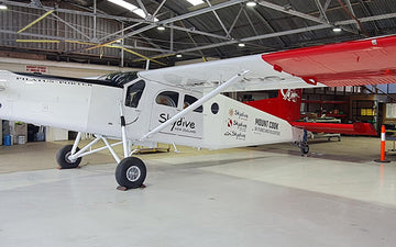 White and red Pilatus Porter aircraft branded with Skydive New Zealand and Skydive Mount Cook logos, parked inside an aircraft hangar.