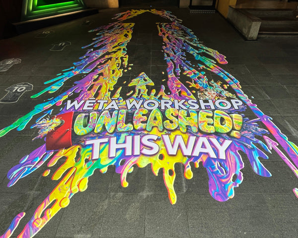 Vibrant 3D-style floor graphic reading “Wētā Workshop Unleashed – This Way,” with neon paint splatters and an explosion of colours guiding visitors.