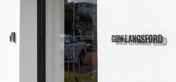 Modern glass entrance with minimalist signage for Gow Langsford Gallery, reflecting parked vehicles and surrounding architecture.