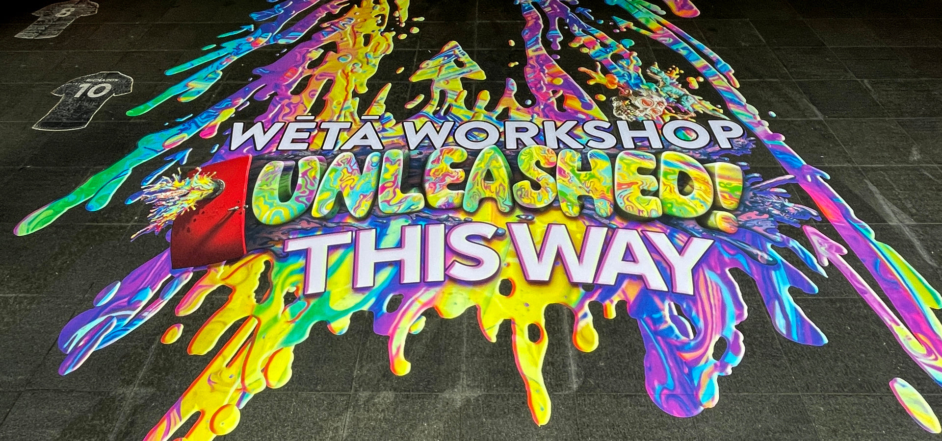 Multicoloured, eye-catching floor decal promoting “Wētā Workshop Unleashed” with dynamic splashes and arrows directing visitors toward the exhibit.