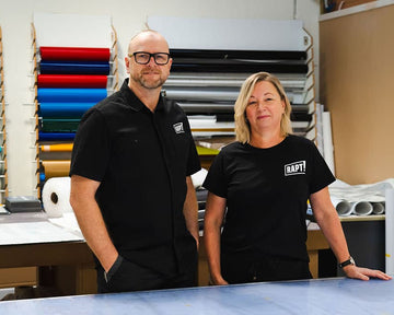 Two people standing in a workshop or studio with rolls of colored material in the background. The person on the left wears a black short-sleeve button-up shirt with a small white logo, and the person on the right wears a black T-shirt with a white 'RAPT' logo.