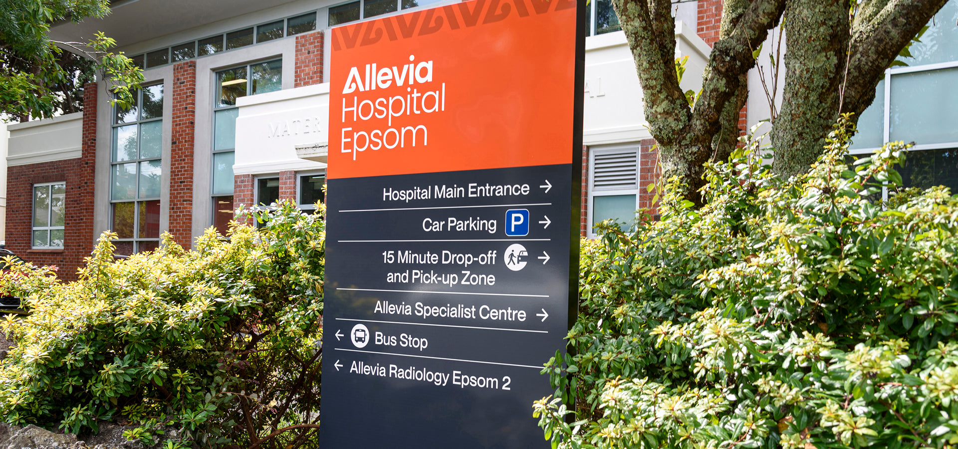 Directional hospital signage outside Allevia Hospital Epsom showing main entrance, parking, drop-off zone, specialist centre, bus stop, and radiology directions.